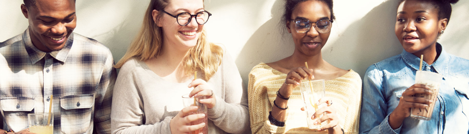 Photo of a diverse group of 4 college students leaning against a wall, laughing and enjoying their various drinks.